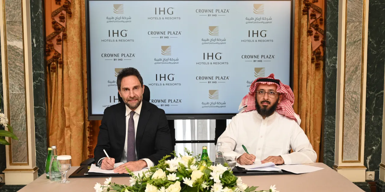 Two executives from IHG Hotels & Resorts and Arbah Tayba Real Estate Company sign an agreement in front of a branded display during a formal ceremony.