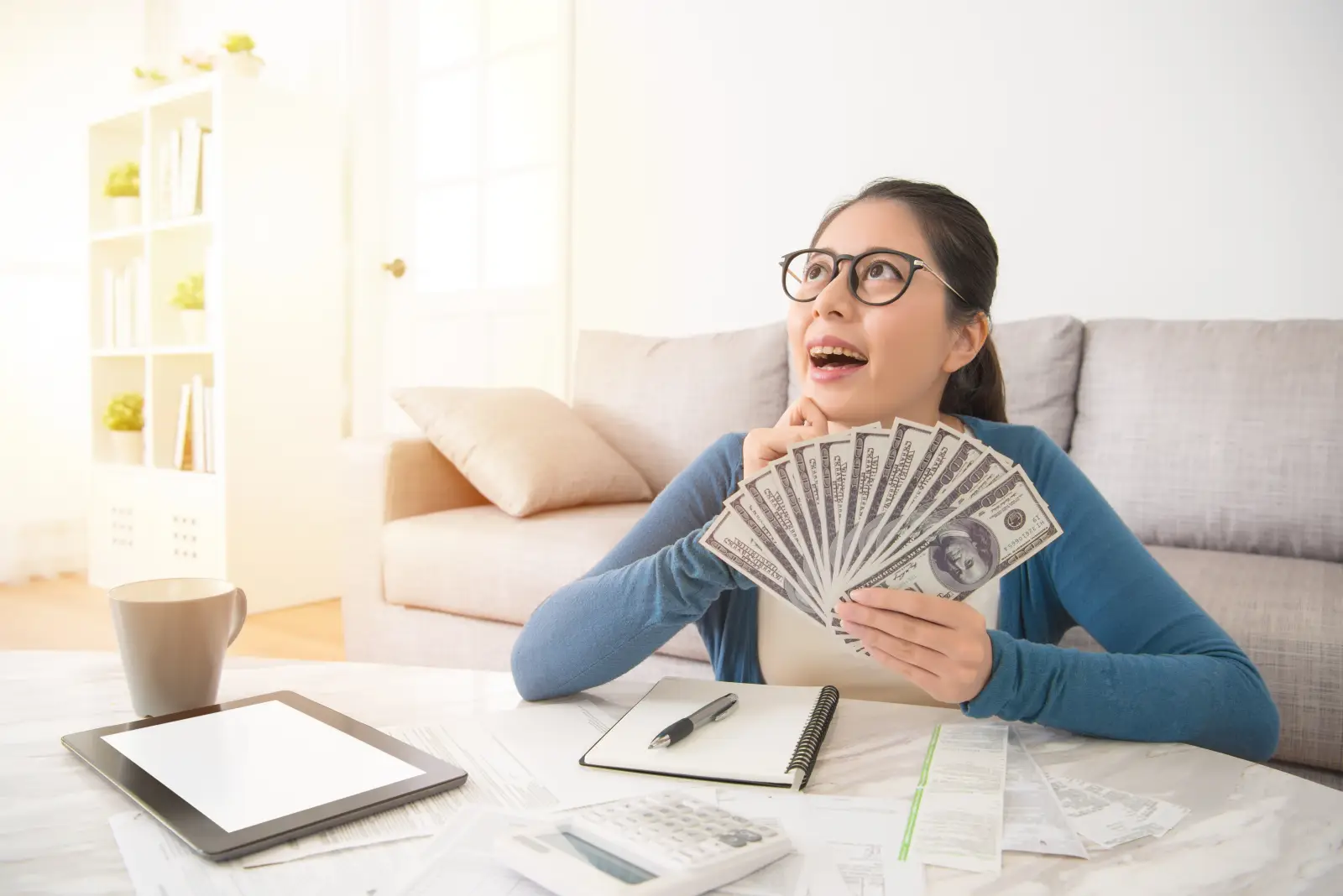 What It Takes To Feel Wealthy In 2025, Survey Reveals 2 Woman smiling while holding cash and planning her budget, feeling wealthy at home.