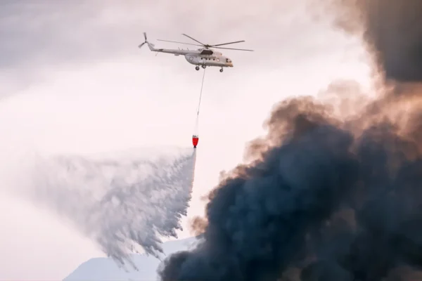 A firefighting helicopter drops water over thick black smoke from a wildfire.