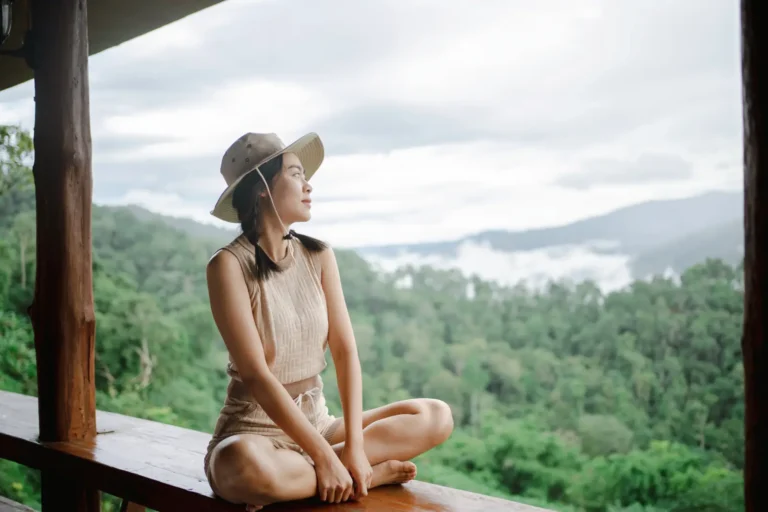 Woman in safari hat sitting cross-legged on a wooden ledge, peacefully enjoying a misty forest and mountain view.