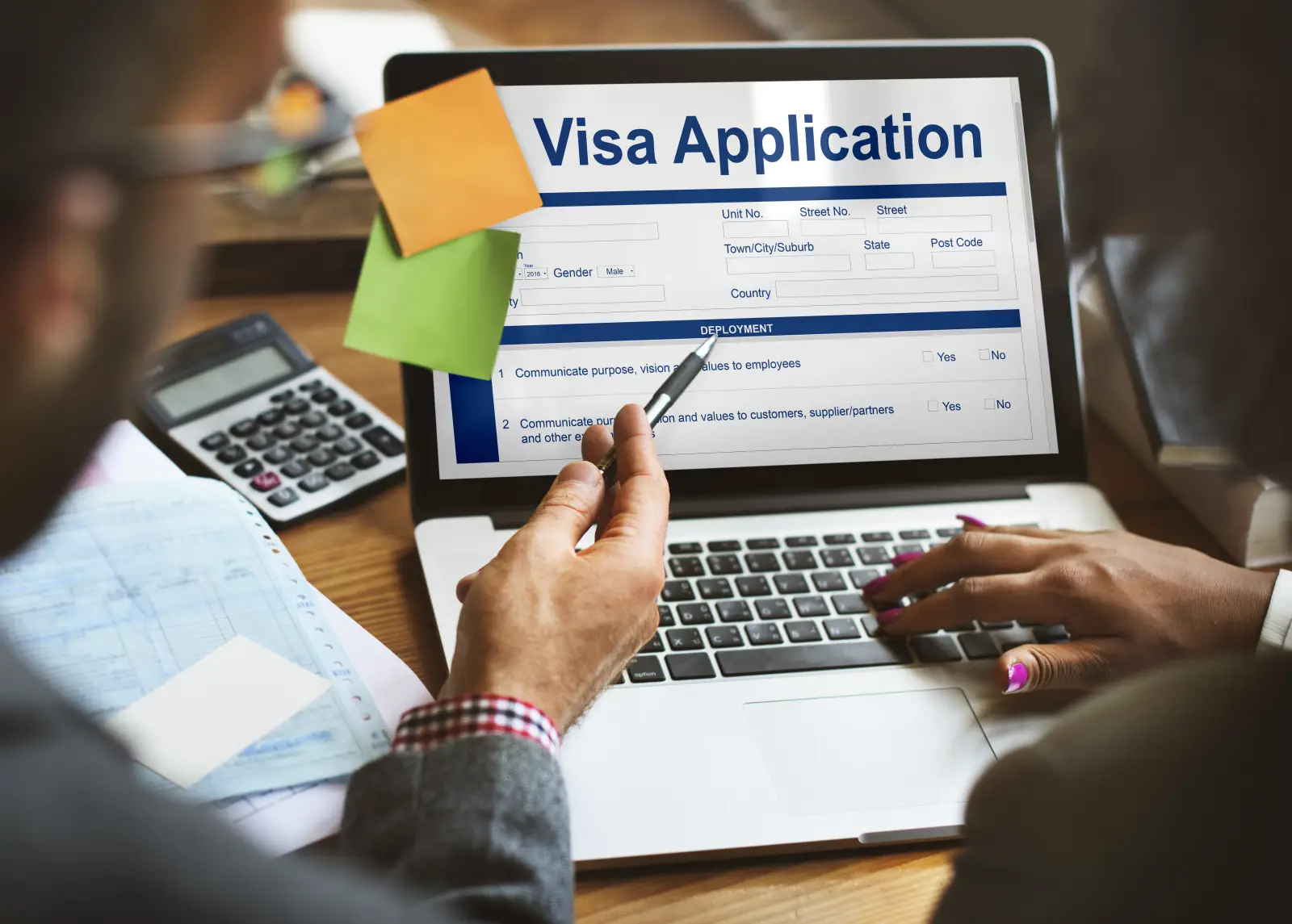 Two people reviewing an online visa application form on a laptop, with documents and a calculator on the desk