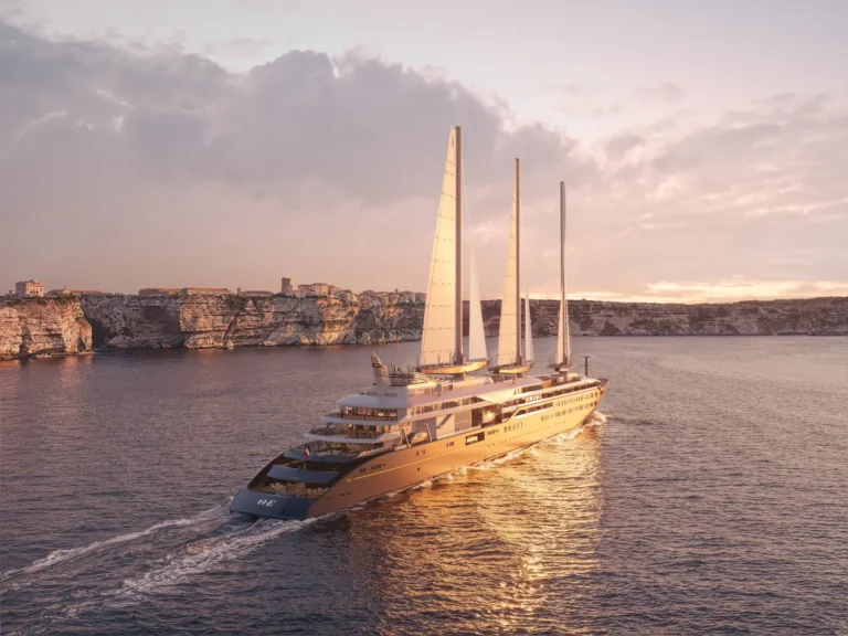Luxury Travel 17 Orient Express Corinthian sailing yacht cruising past rocky cliffs at sunset with its three sails upright.
