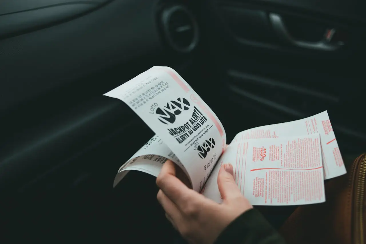 Thai Lottery On Holiday: What Tourists Need To Know Before Playing Online 2 Person holding Lotto Max lottery tickets inside a car, reviewing the printed slips with jackpot alert visible.