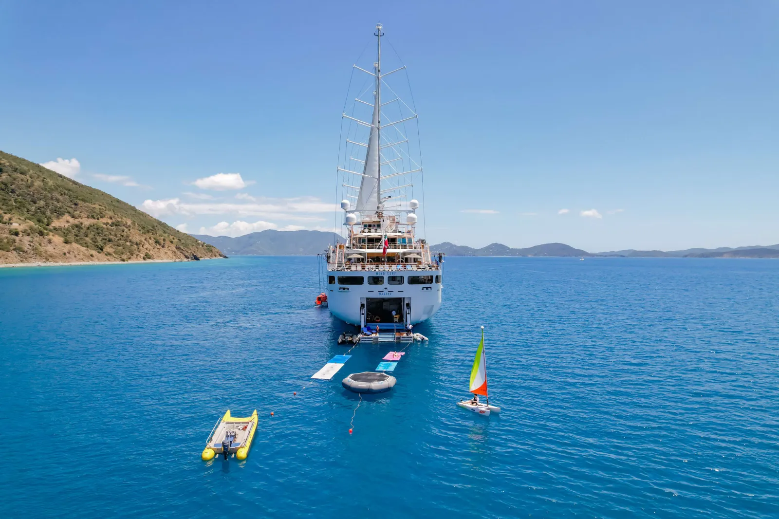 Windstar Cruises Adds Free All-Inclusive Upgrade 3 Windstar Cruises sailing yacht anchored in a Caribbean bay with inflatable platforms, a small sailboat, and a motorized tender floating in clear blue water