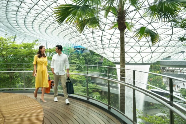 A couple walks along an indoor garden walkway at Jewel Changi Airport in Singapore, with lush greenery and the Rain Vortex waterfall in the background.