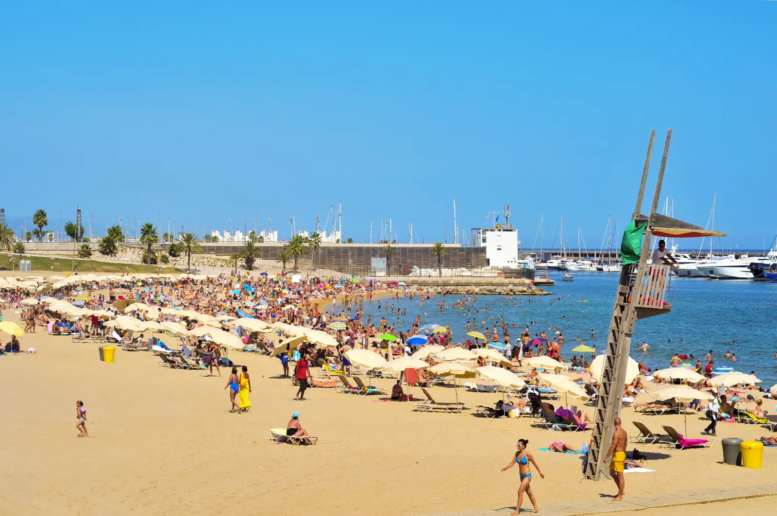 Crowded summer day at Barceloneta Beach in Barcelona with sunbathers, umbrellas, a lifeguard tower, and yachts in the background.