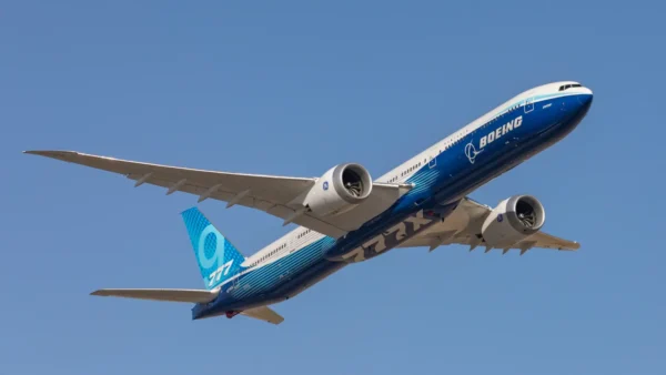 Home 3 Boeing 777-9 aircraft in flight against a clear blue sky, showing its extended wingspan and twin-engine configuration.