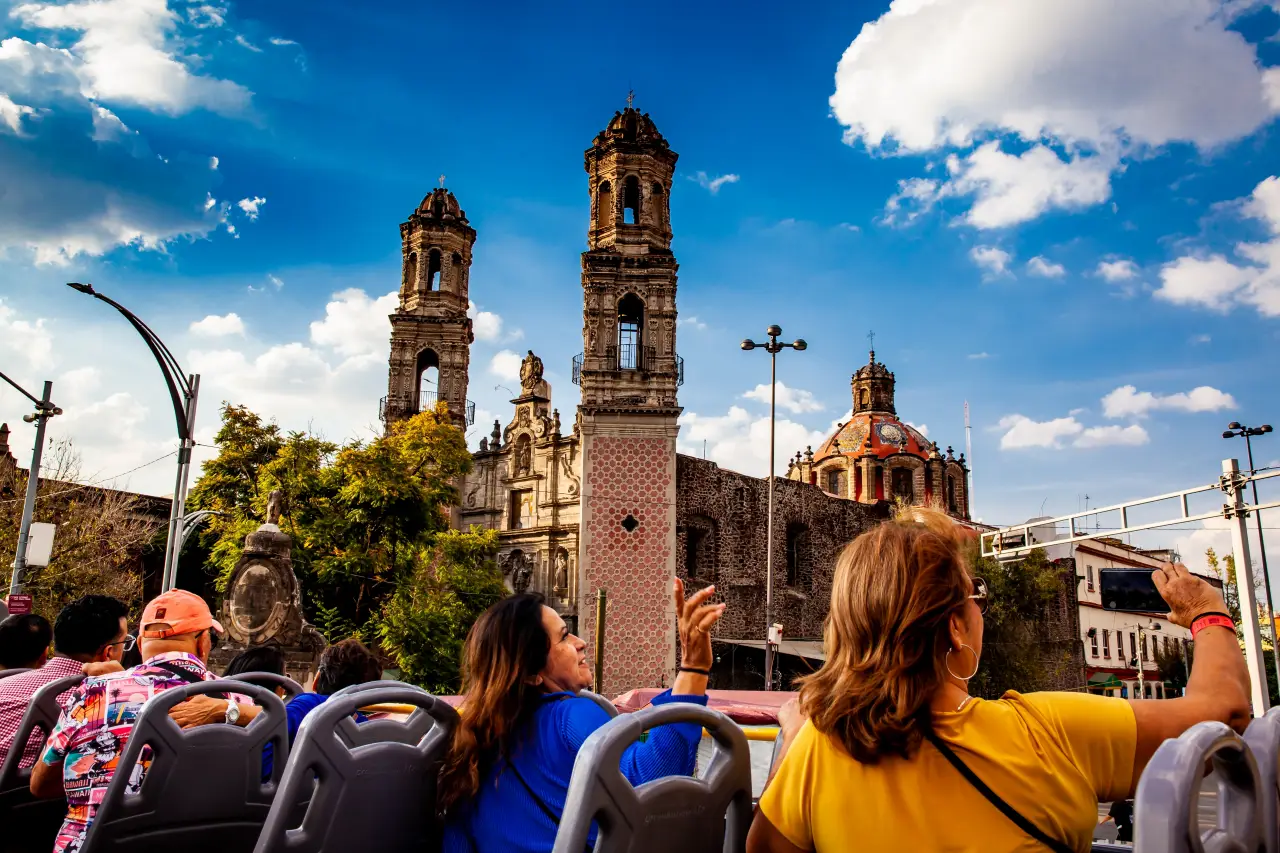Tourists on an open-top bus in Mexico City admire historic colonial architecture with church towers under a bright blue sky.