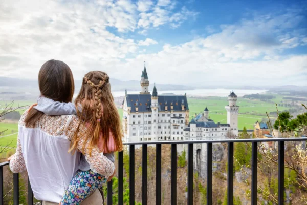 Mother and daughter standing on a viewpoint, admiring Neuschwanstein Castle in Bavaria, Germany, with green fields and mountains in the background.