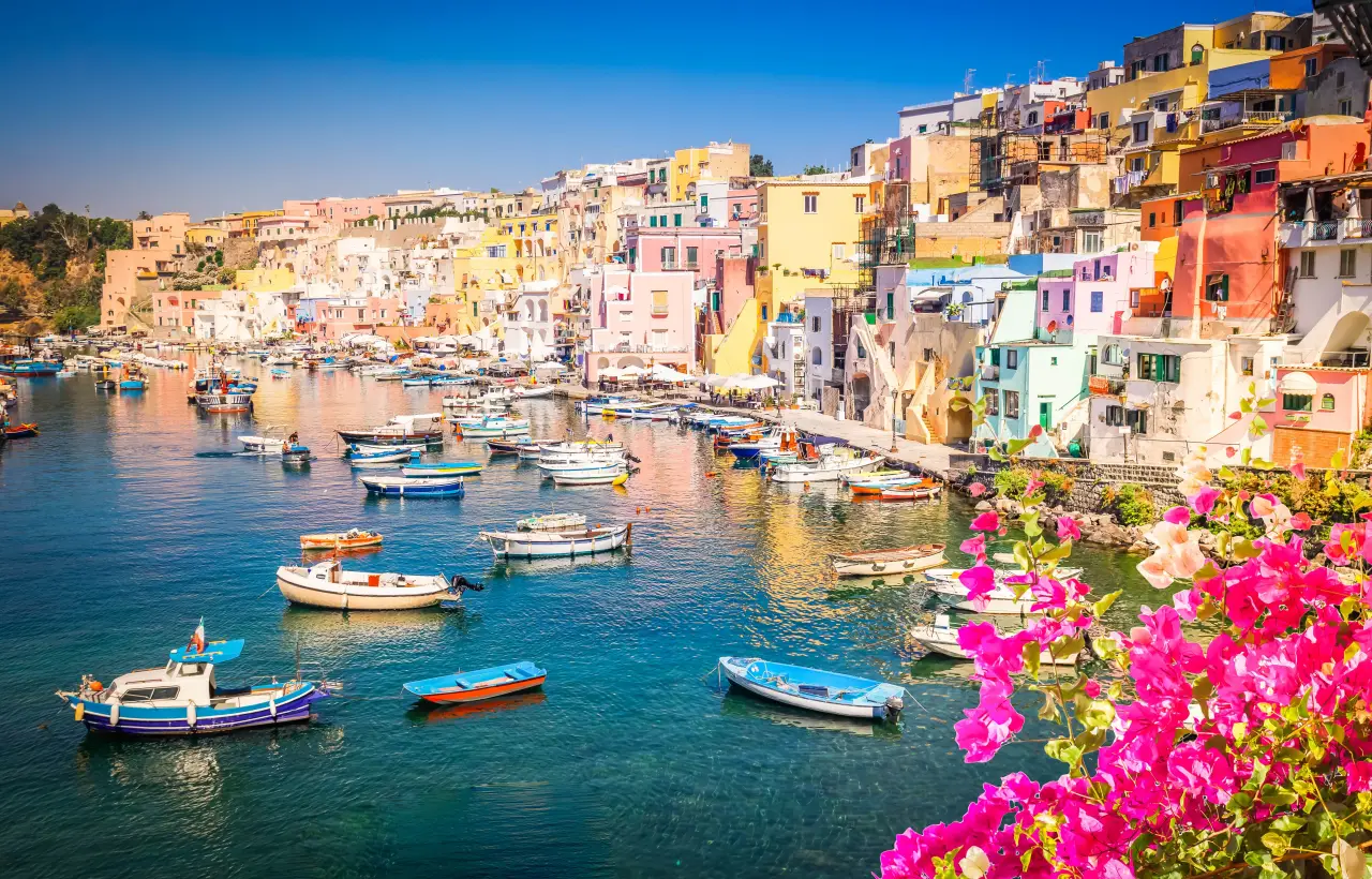 Why Procida Is Italy’s Most Authentic Island Escape 2 Colorful fishing village of Marina Corricella on Procida Island, Italy, with pastel houses, boats in the harbor, and bright pink flowers in the foreground.