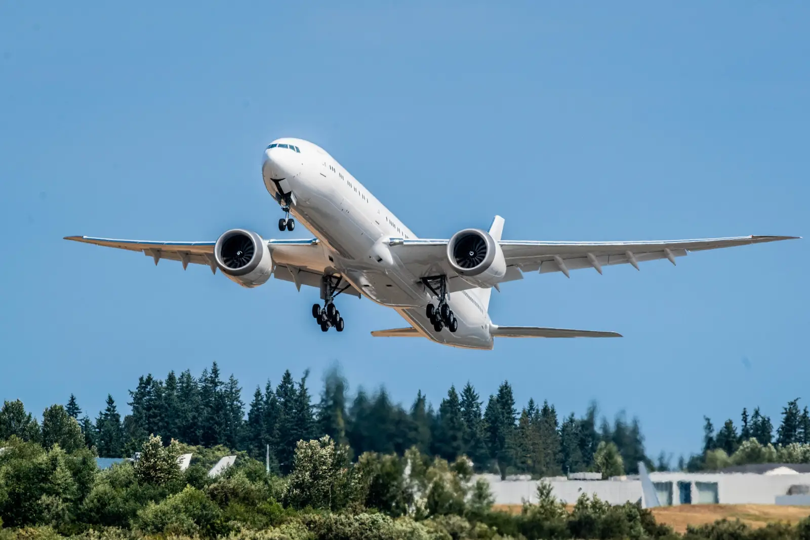 All-white Boeing 777X aircraft taking off during a test flight in Seattle, with landing gear extended and forest in the background.