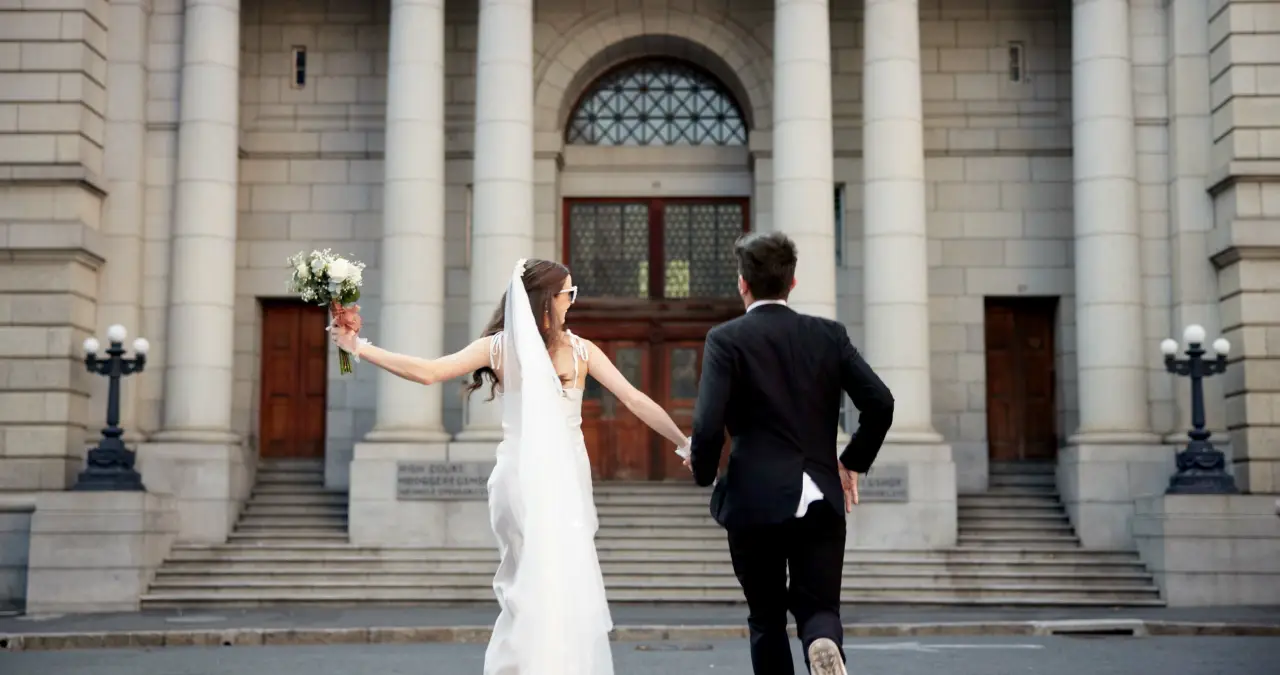 Smart Ways To Save For Your 2026 Wedding 2 Bride and groom holding hands and running up the steps of a historic stone building with tall columns, the bride holding a bouquet in celebration.