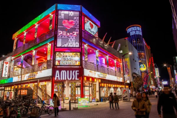 Home 4 A brightly lit entertainment and nightlife district in Japan at night, featuring colorful neon signs, a building labeled “Amuse,” and people walking along the street.