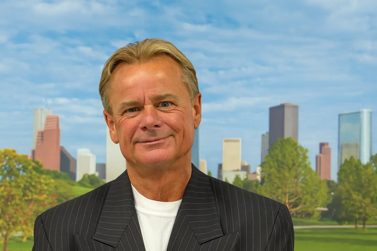 Portrait of Jordy Tollett smiling in front of the Houston skyline with trees and skyscrapers in the background.