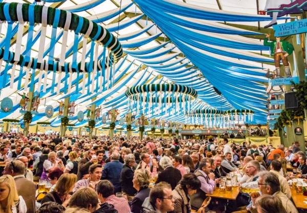 Large beer tent at Oktoberfest in Munich filled with people enjoying drinks and food, decorated with blue and white drapes and festive wreaths.