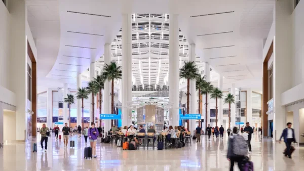 Interior of Orlando International Airport’s modern terminal with palm trees, travelers walking with luggage, and people seated in a central waiting area.