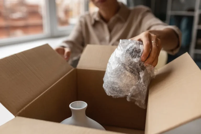 Person carefully packing a fragile ceramic vase into a cardboard box using bubble wrap for protection.
