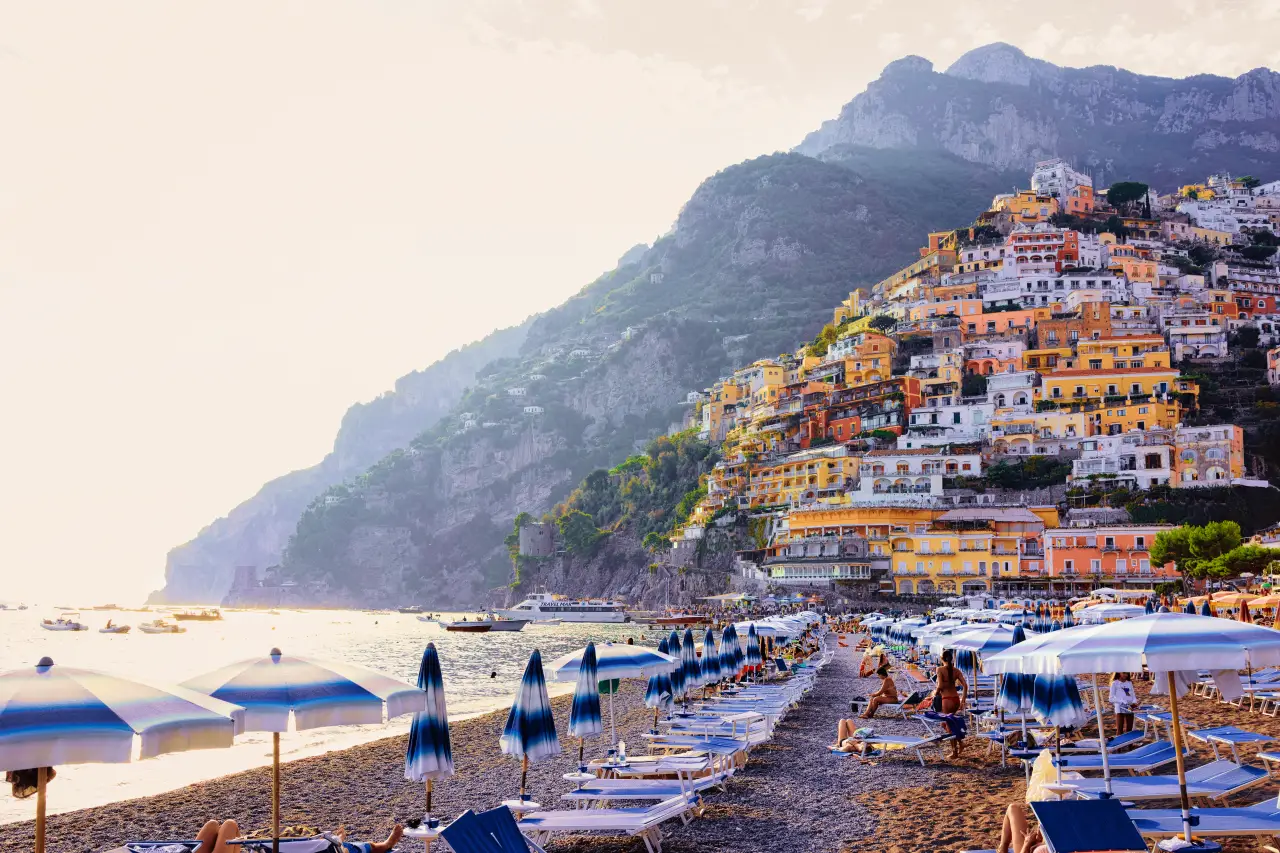 Italian Beaches See Sharp Drop In Visitors This Summer 2 View of Positano Beach on the Amalfi Coast, Italy, with rows of umbrellas and sunbeds along the shoreline and colorful hillside houses above the sea.