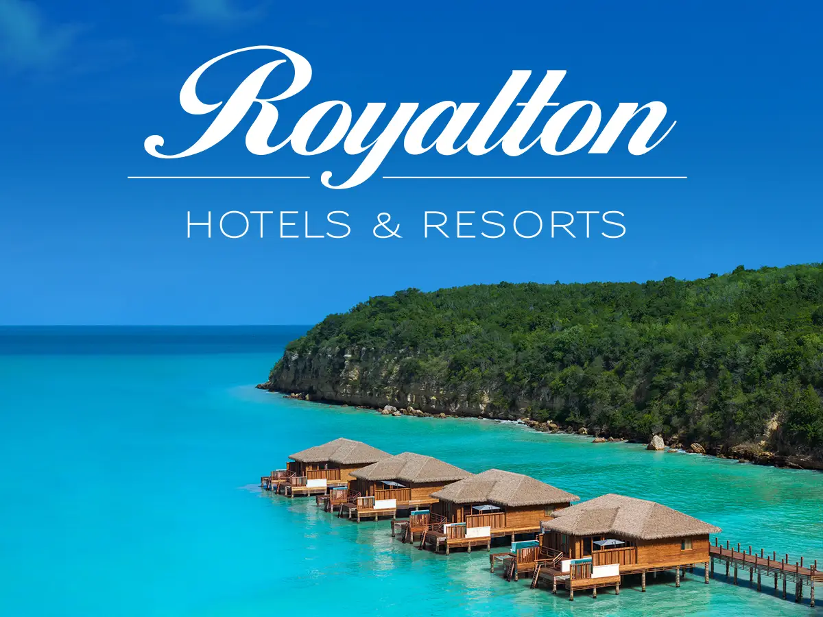 Overwater villas at a Royalton Hotels & Resorts property in the Caribbean, surrounded by turquoise sea and lush green cliffs.