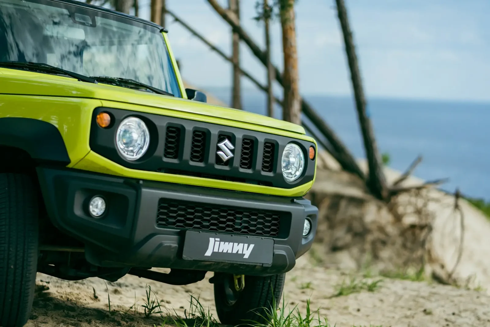 A bright green Suzuki Jimny parked on sandy terrain near the coast, highlighting its rugged design and off-road capability.