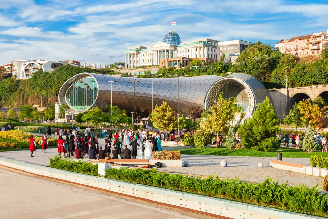 Top Wedding Locations In Georgia 2 A lively gathering in Rike Park, Tbilisi, Georgia, with modern architecture, green gardens, and the Presidential Palace in the background.