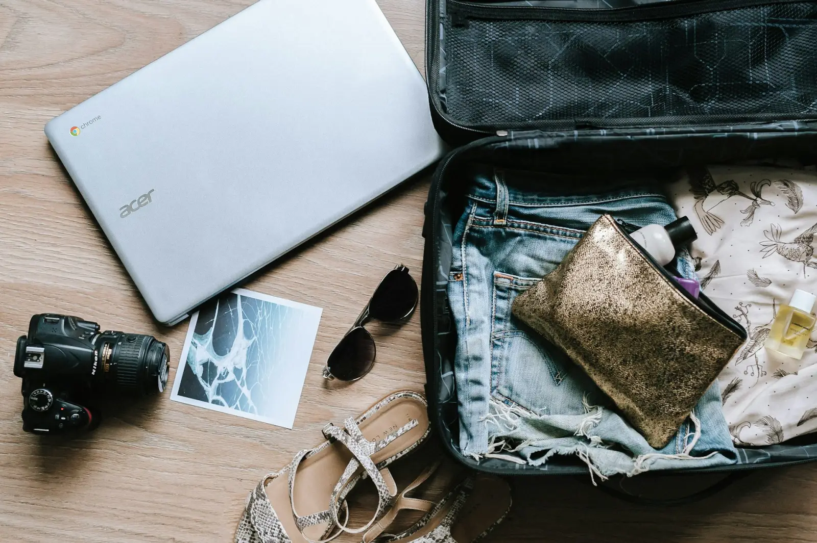 Open suitcase with summer clothes, sandals, sunglasses, camera, and a laptop on a wooden floor, ready for travel packing.