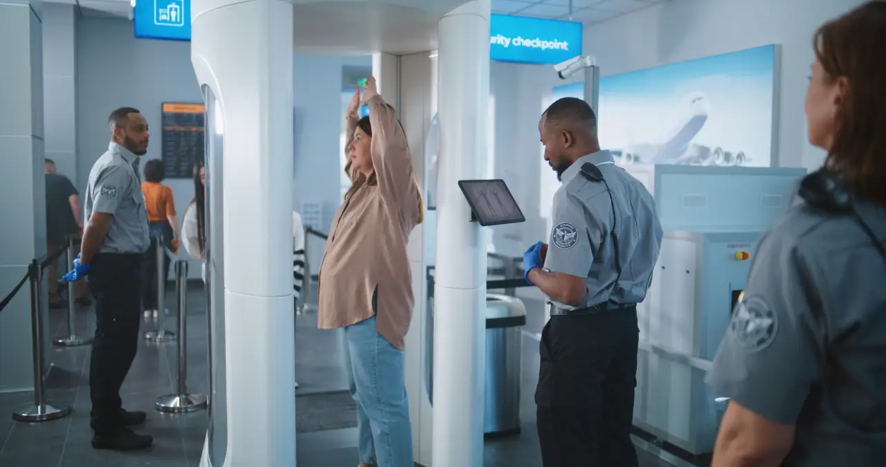 TSA Airport security officers monitor a passenger undergoing a body scanner check at a security checkpoint, with screening equipment and travelers in the background.