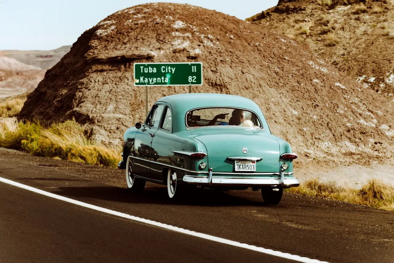Vintage teal car driving along a desert highway toward Tuba City and Kayenta, with rocky hills in the background.