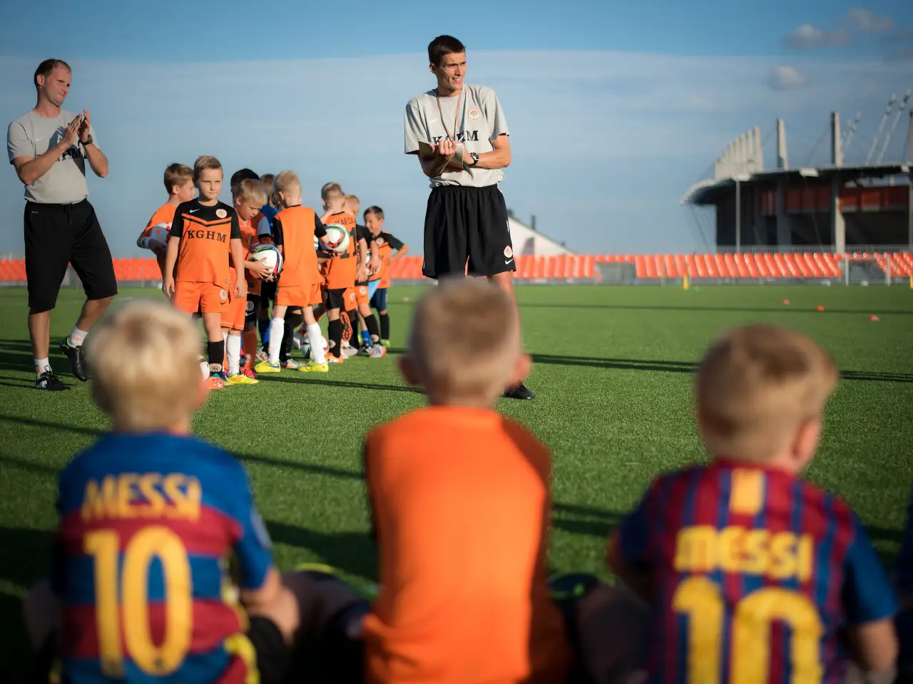 Children in bright football kits gather on a pitch during a youth training session, guided by coaches under a sunny sky at a stadium.