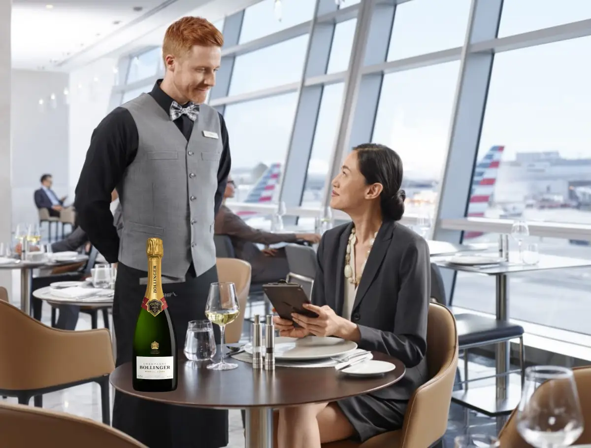 Waiter serving a female passenger Bollinger Champagne at an American Airlines lounge with planes visible outside the window.