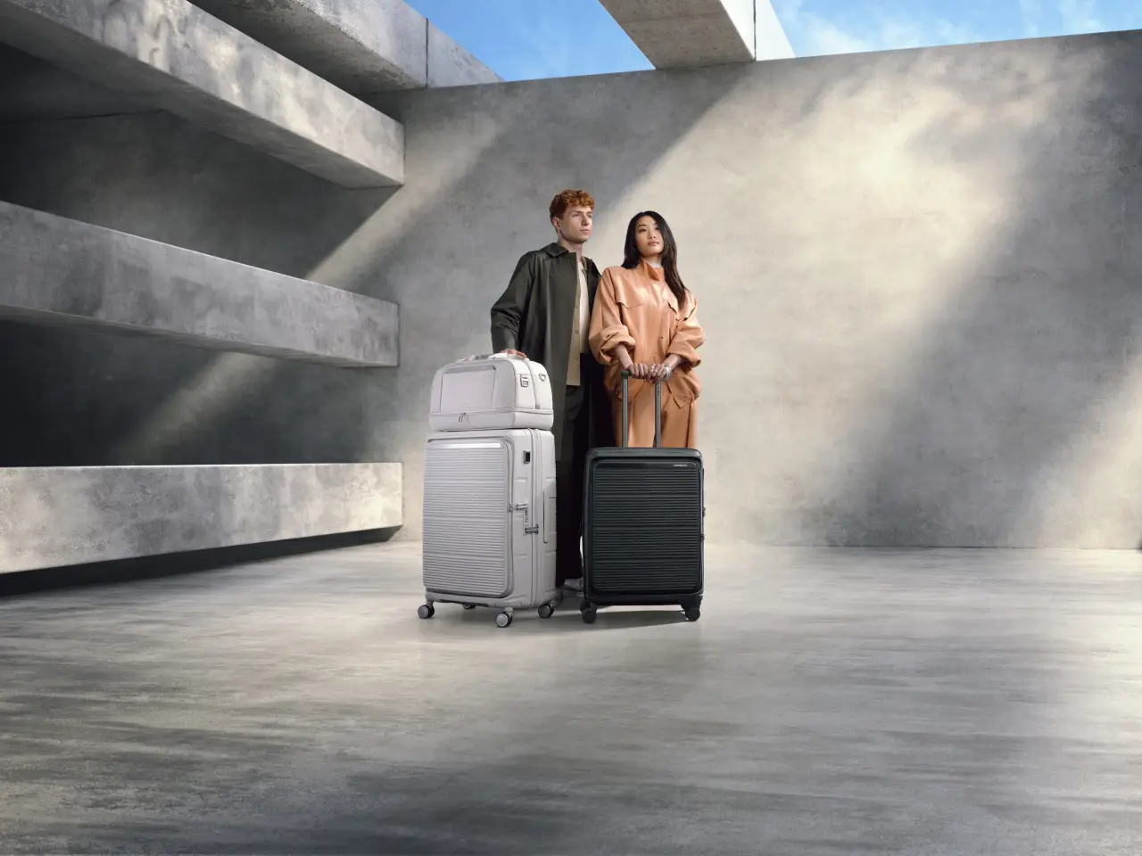 Samsonite Unveils Modular, Eco-Friendly Paralux Luggage For Global Travelers 2 Two models stand in a minimalist concrete space with Samsonite Paralux suitcases, showcasing modular, eco-friendly travel gear in black and white.