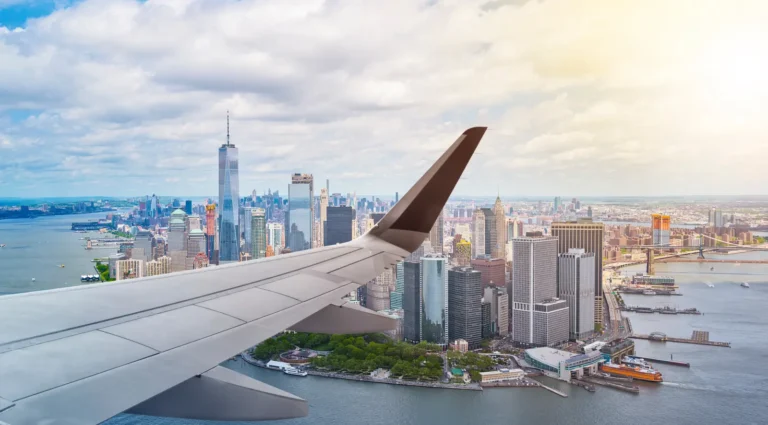 Airplane wing in view while approaching New York City, with the Manhattan skyline and One World Trade Center visible in the background.