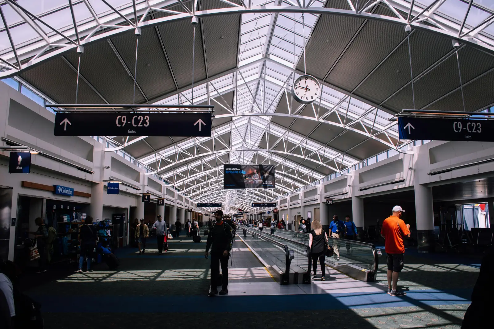 Interior view of a busy airport terminal with skylight ceiling, passengers walking, and signs for gates C9 to C23.