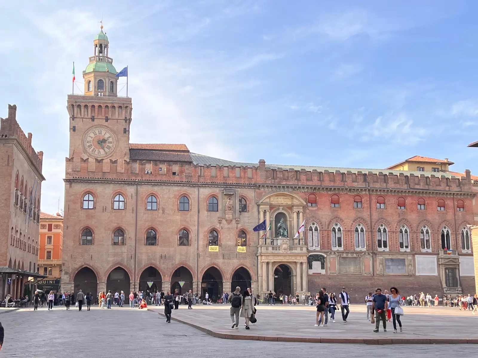 Historic brick building with clock tower and arched windows in Bologna’s main square, people walking in the foreground
