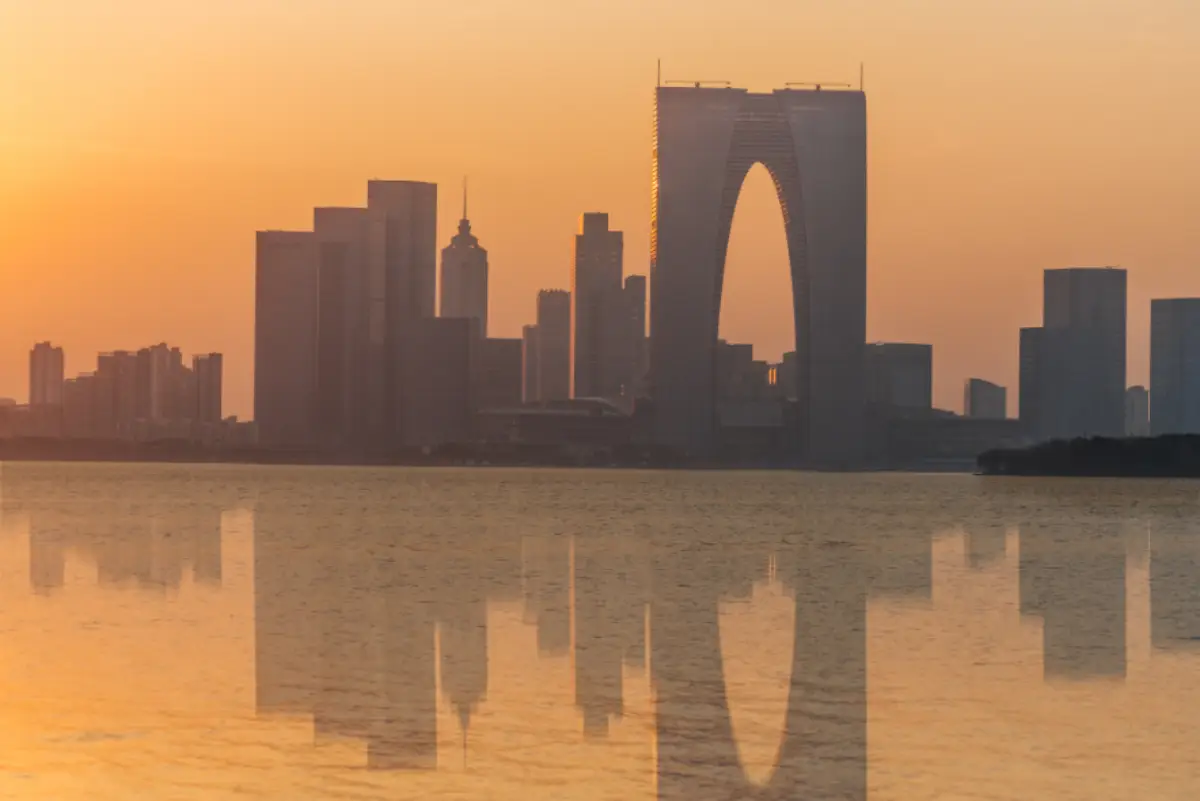 Sunset skyline with modern skyscrapers and a landmark arch-shaped tower reflected on the water.