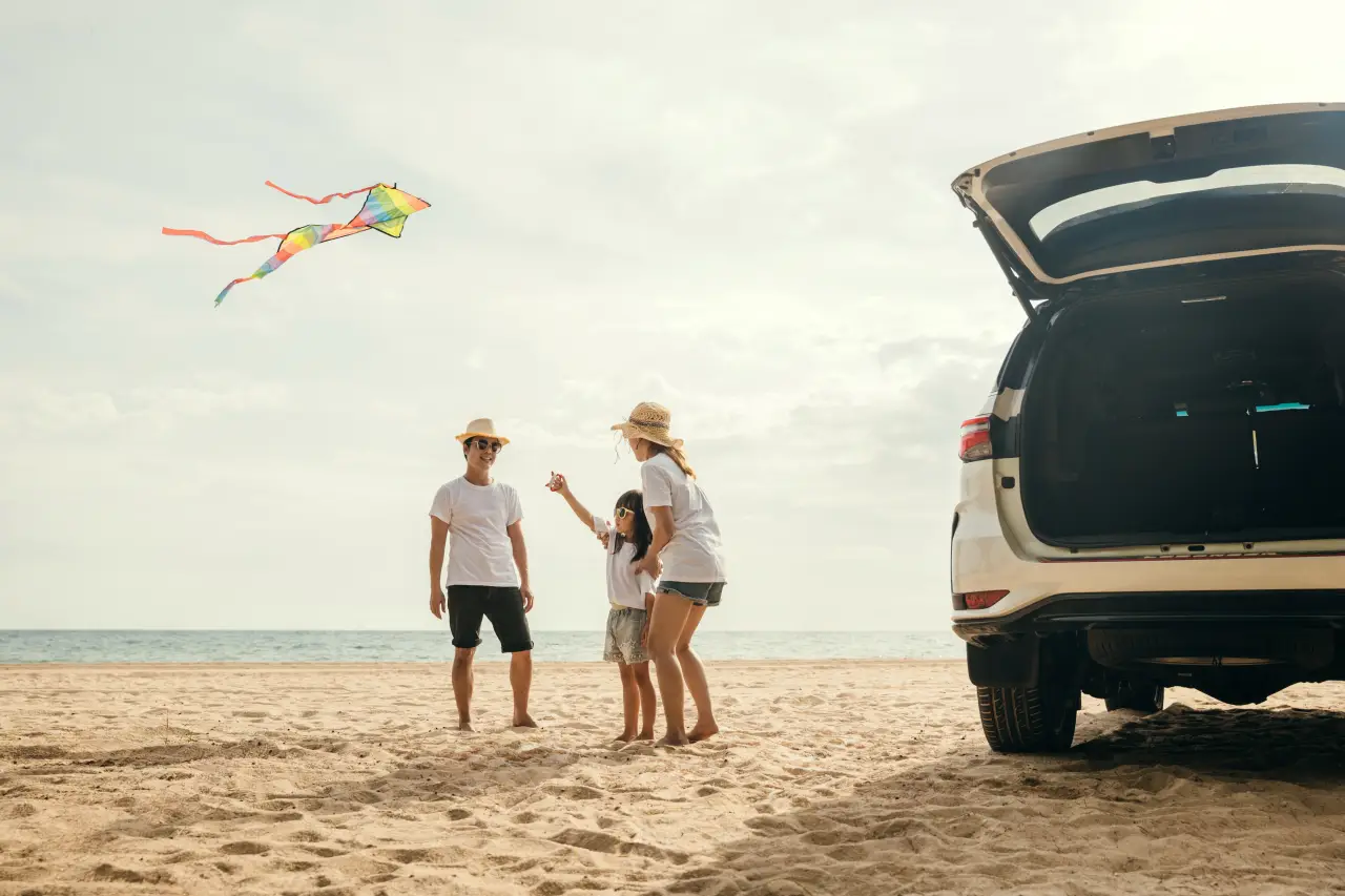 Family flying a kite on the beach near a parked car with open trunk