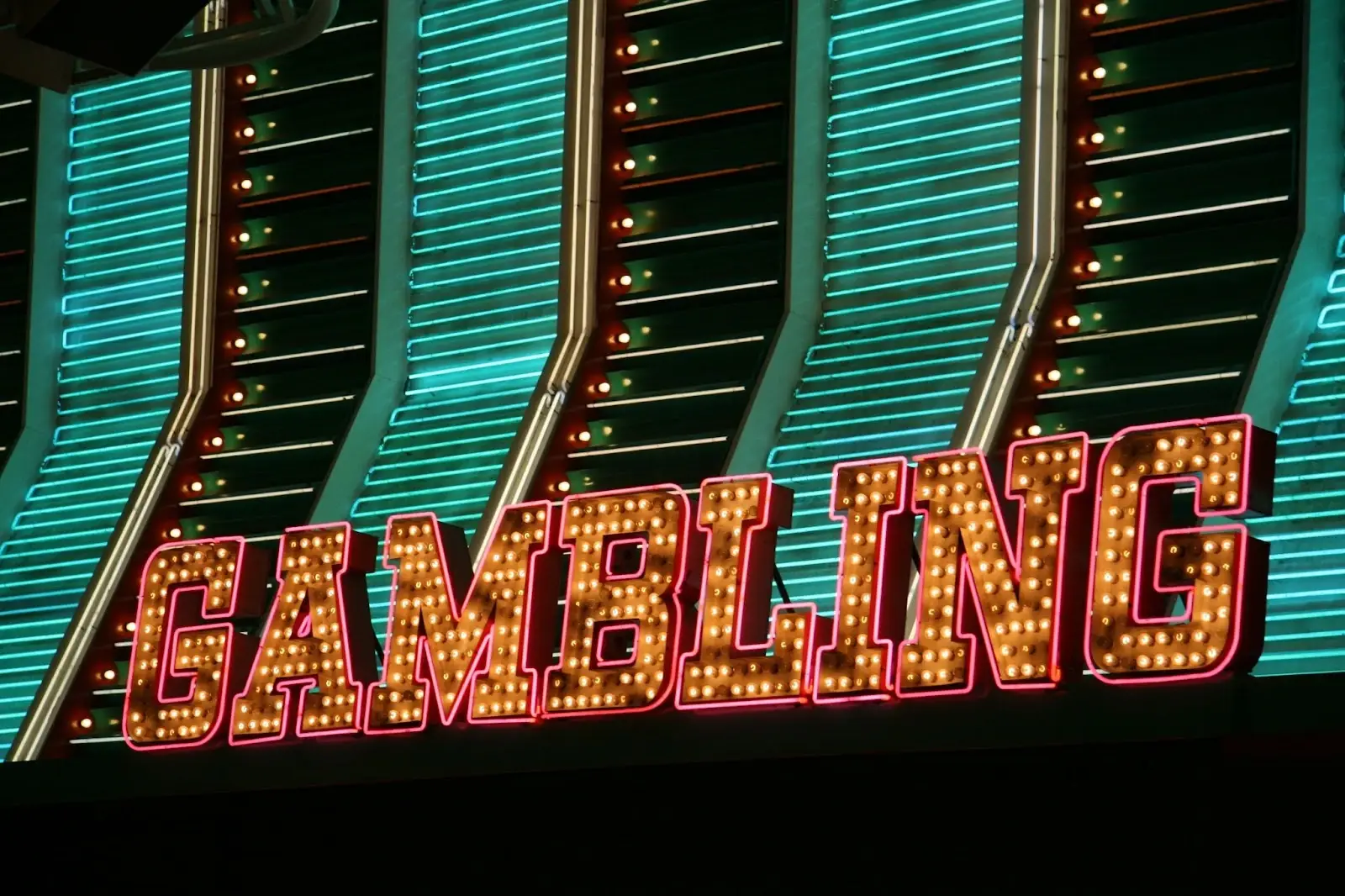 Close-up of a neon “GAMBLING” sign lit with marquee-style bulbs against a teal background.