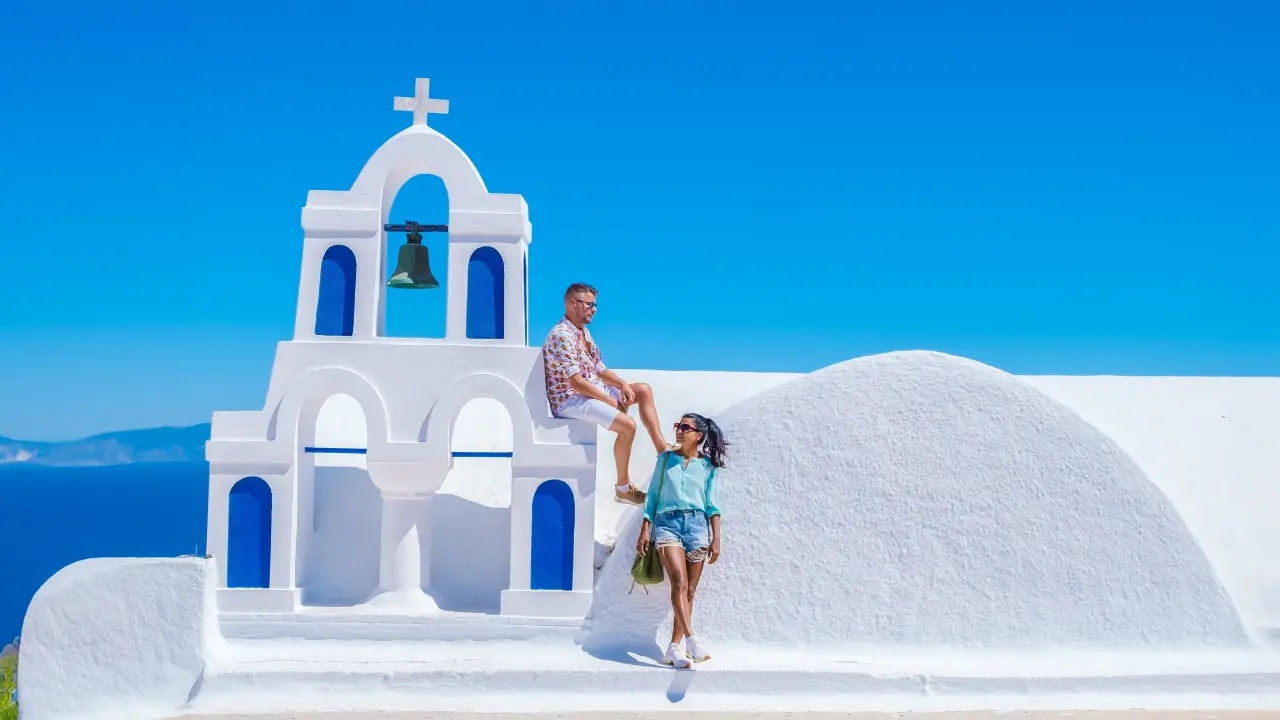 Couple enjoying a sunny day by a whitewashed chapel with blue accents on a Greek island.