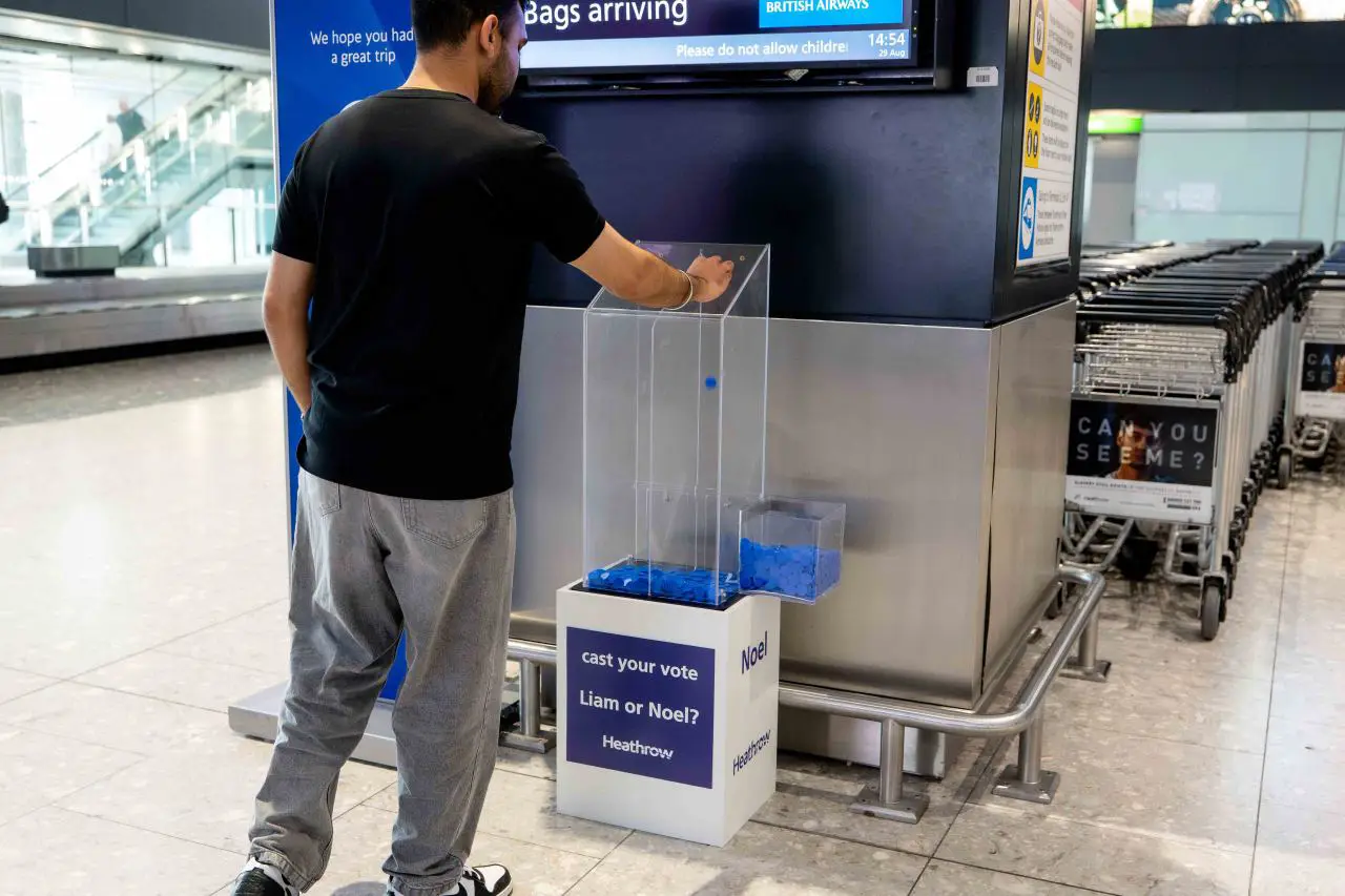 Heathrow Launches Oasis Fan Vote Ahead Of Final London Reunion Shows 3 Passenger casts a vote at Heathrow Airport in a Team Liam vs Team Noel Oasis fan poll display.