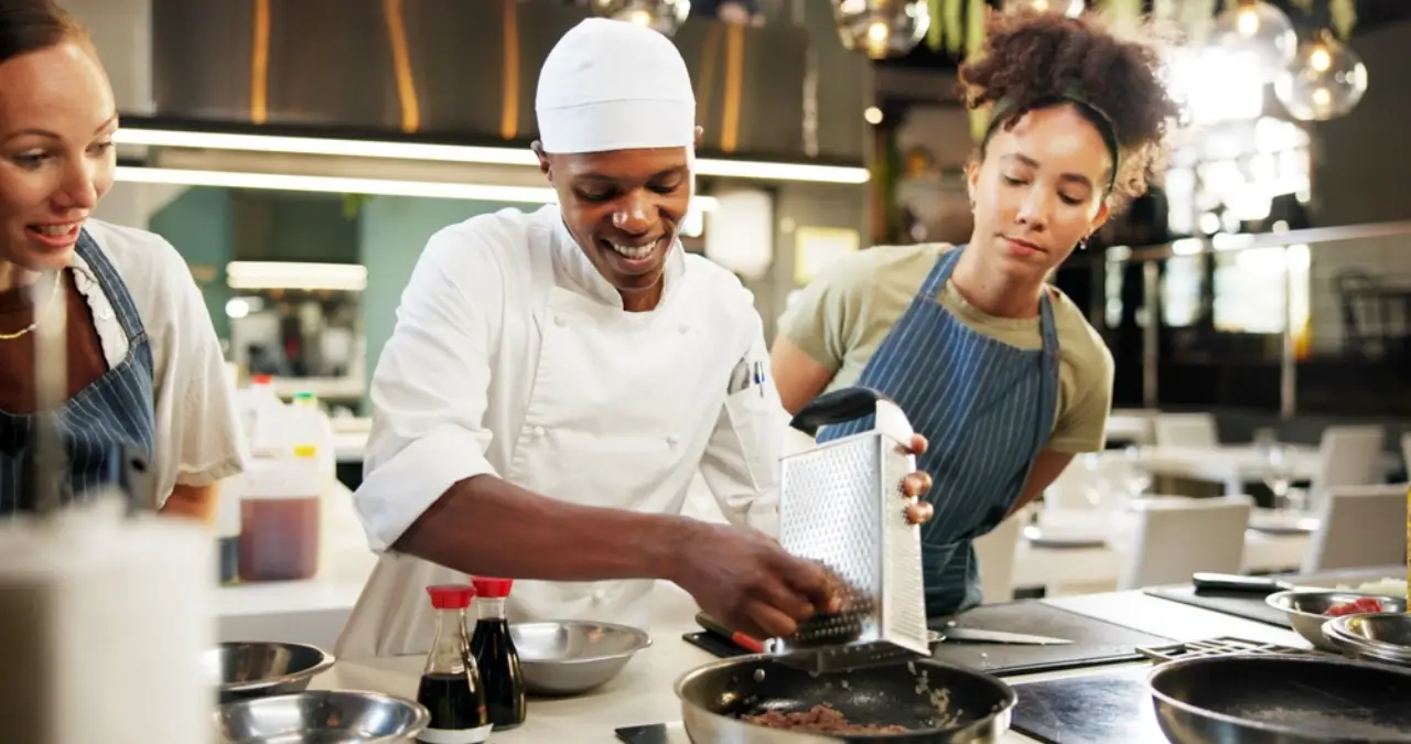 Hospitality instructor in chef uniform grating food while smiling with two students during a culinary training session in a professional kitchen.