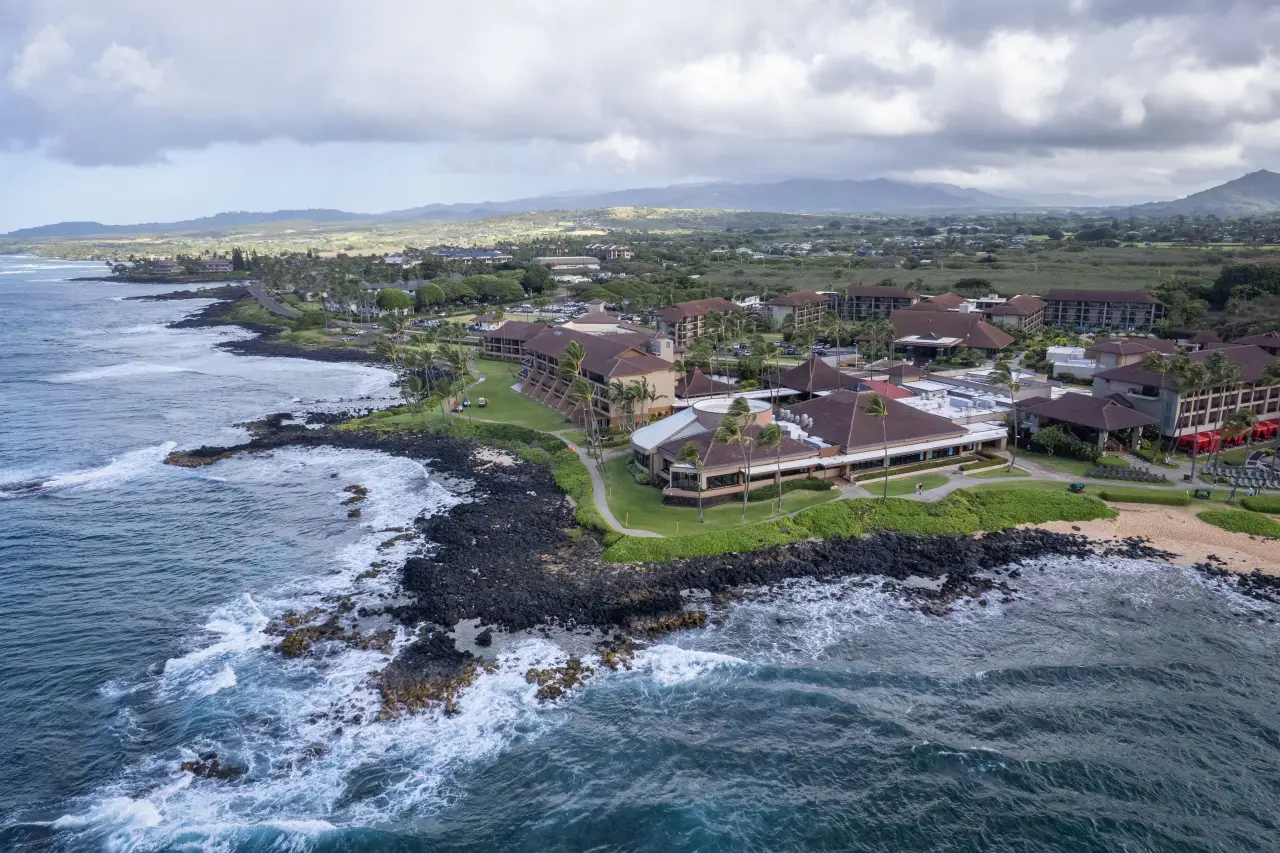 Aerial view of a coastal resort with lush greenery and ocean waves in Kaua‘i, Hawai‘i under a cloudy sky.