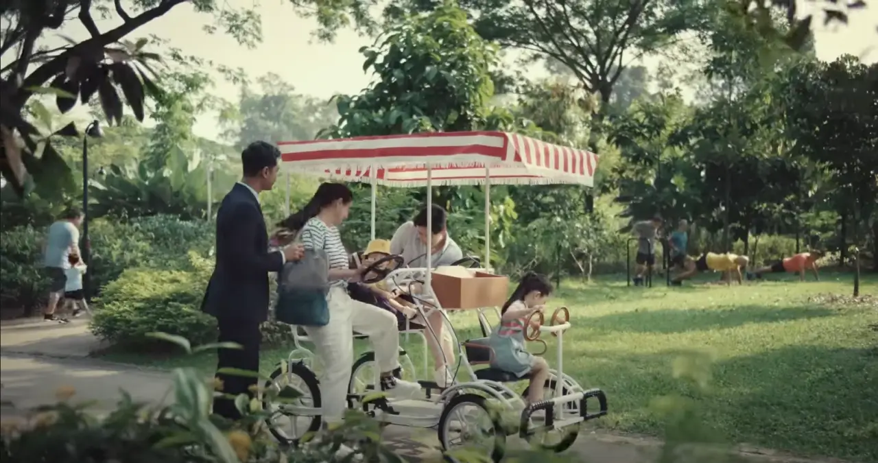 A family rides a quad bicycle in a lush Singapore park, assisted by a Singapore Airlines cabin crew member during a scene from an in-flight safety video.