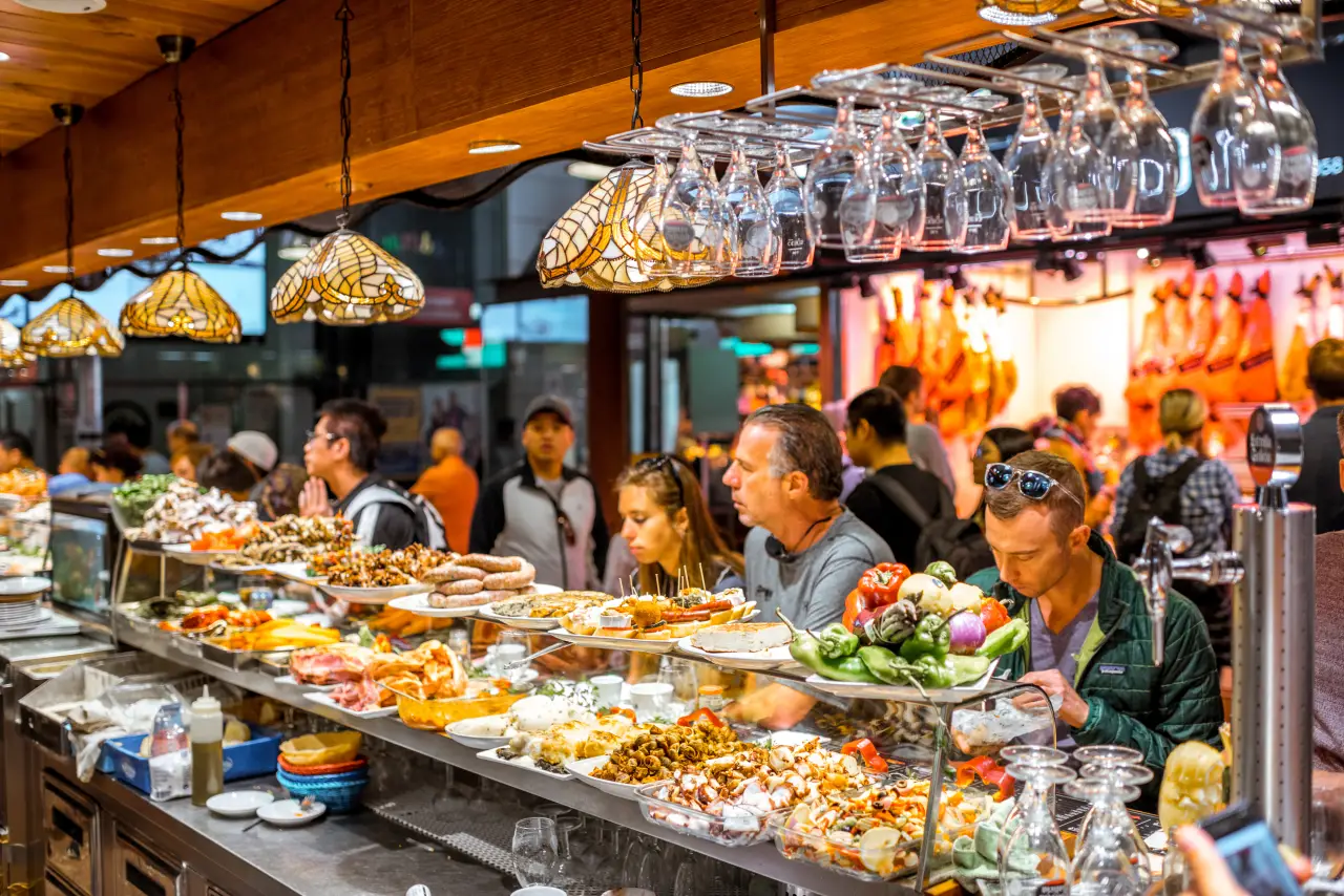 People dining at a busy Spanish tapas bar with assorted dishes and hanging wine glasses