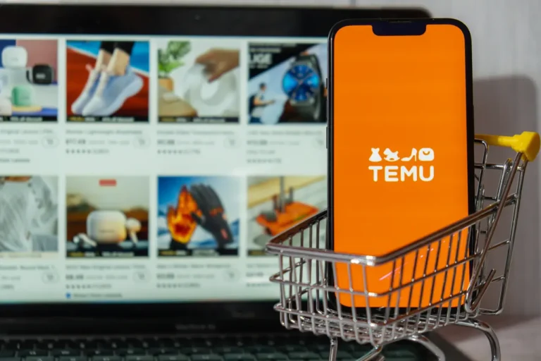 A smartphone with the Temu logo sits in a miniature shopping cart in front of a laptop displaying the Temu online marketplace.