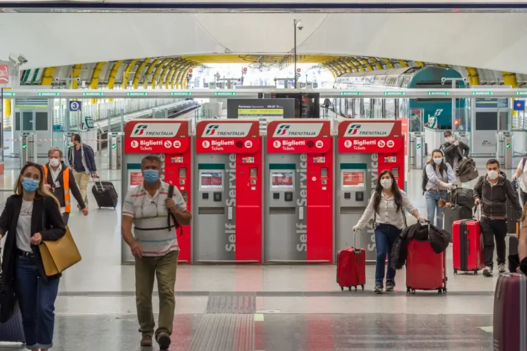 Travelers wearing face masks walk past Trenitalia ticket machines inside a train station in Italy.