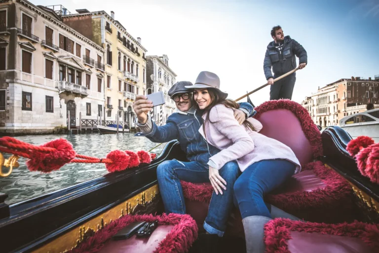 Couple taking a selfie while riding a gondola in Venice with gondolier steering on a canal.