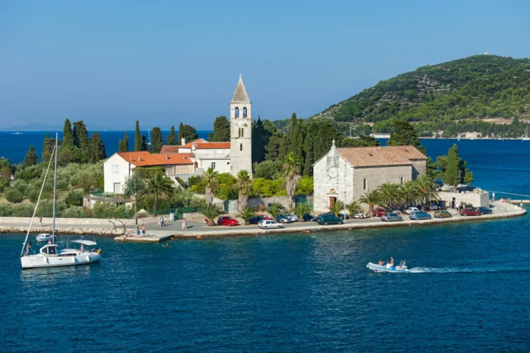 St. Juraj church on Vis Island, Croatia, with stone bell tower, palm trees, and boats in the Adriatic Sea.