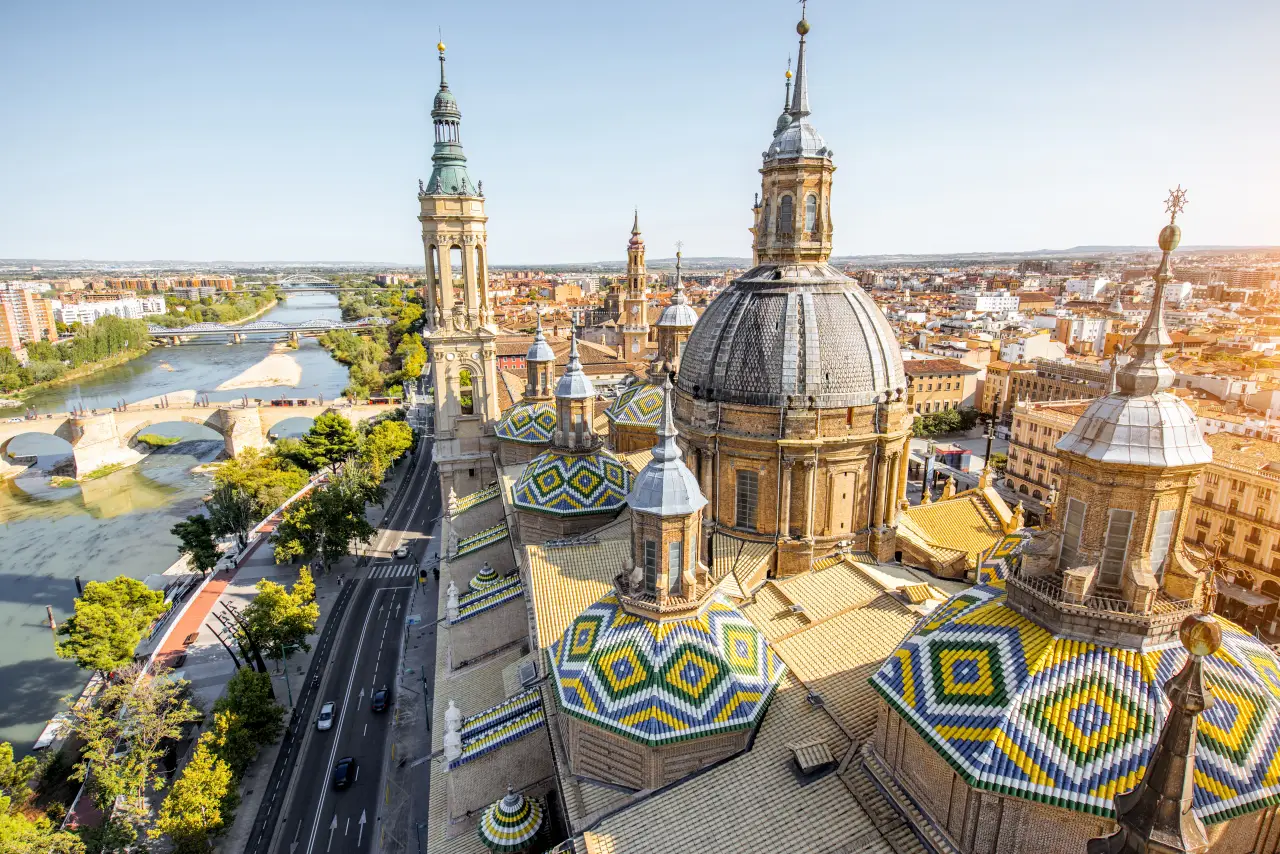 Aerial view of Zaragoza, Spain, featuring the Basilica of Our Lady of the Pillar with its colorful tiled domes and the Ebro River in the background.
