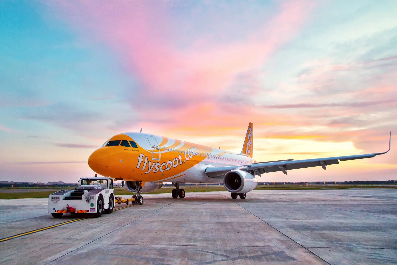 Scoot Airlines aircraft on airport runway at sunset with pink and orange sky.