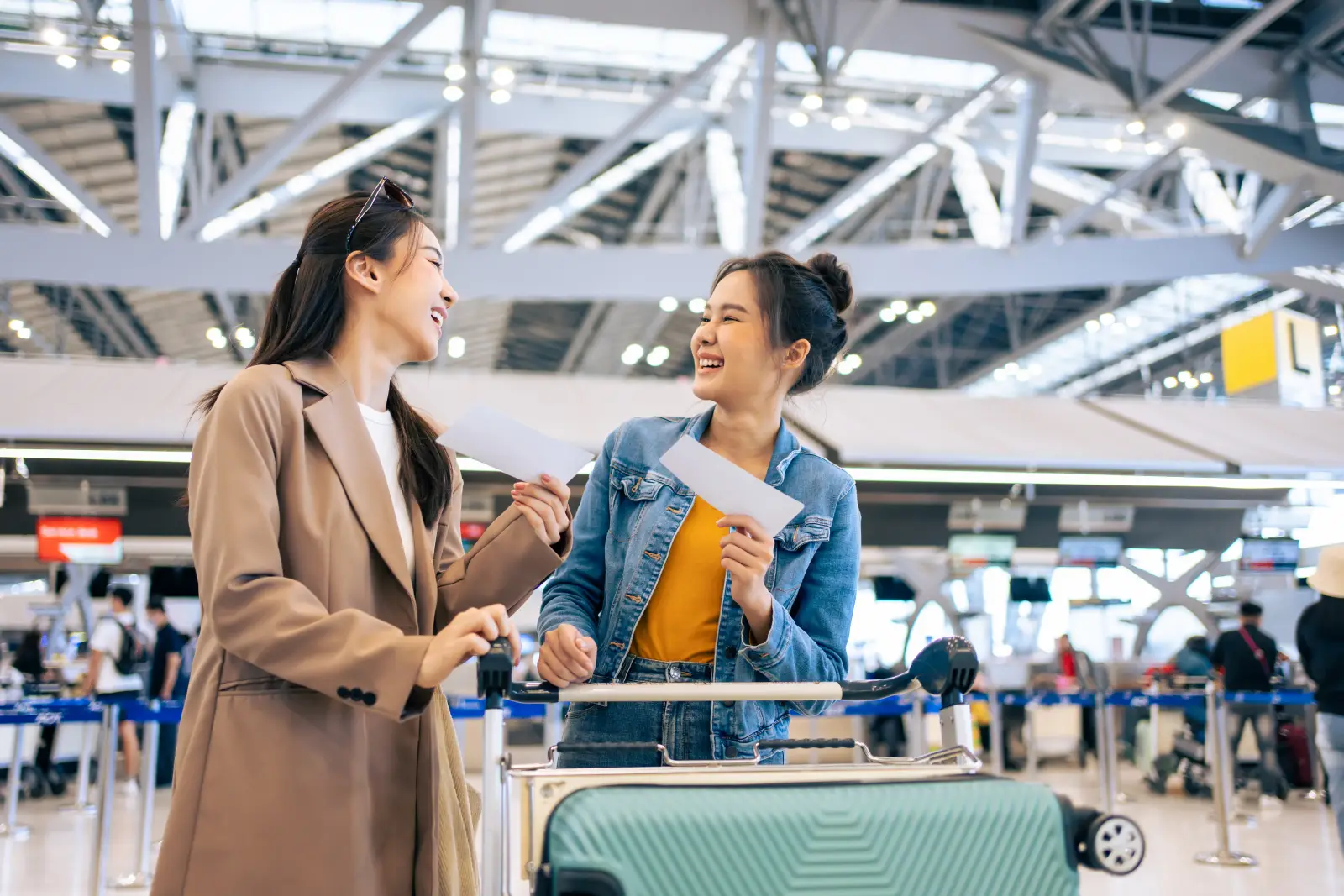 Two travelers share a laugh while holding boarding passes at a busy airport check-in area.